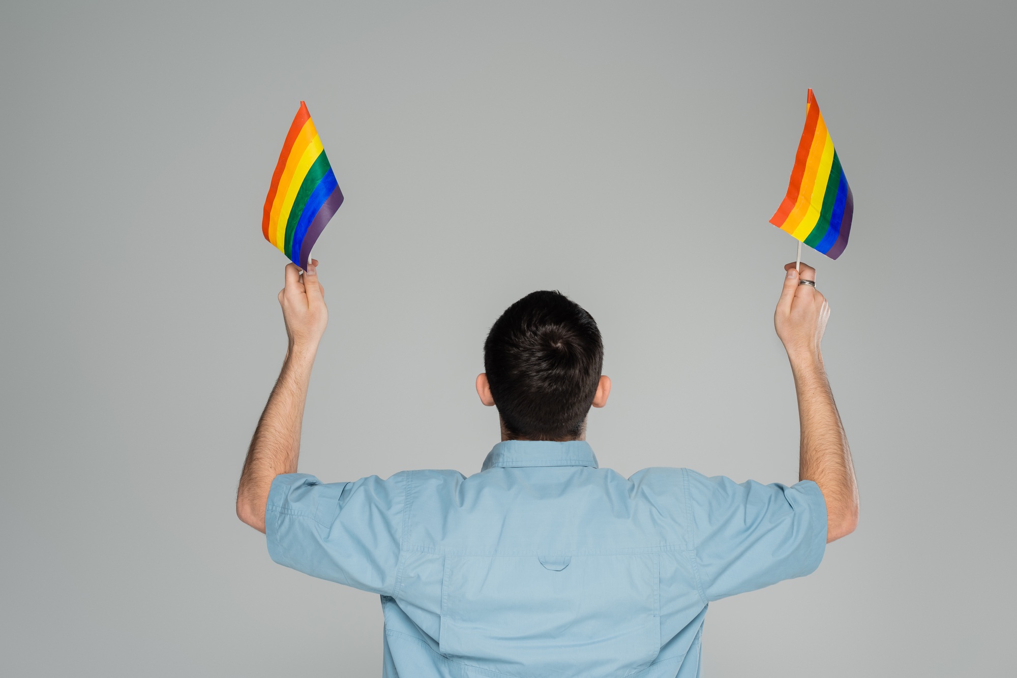Back view of brunette gay man holding lgbt flags isolated on grey, International Day Against